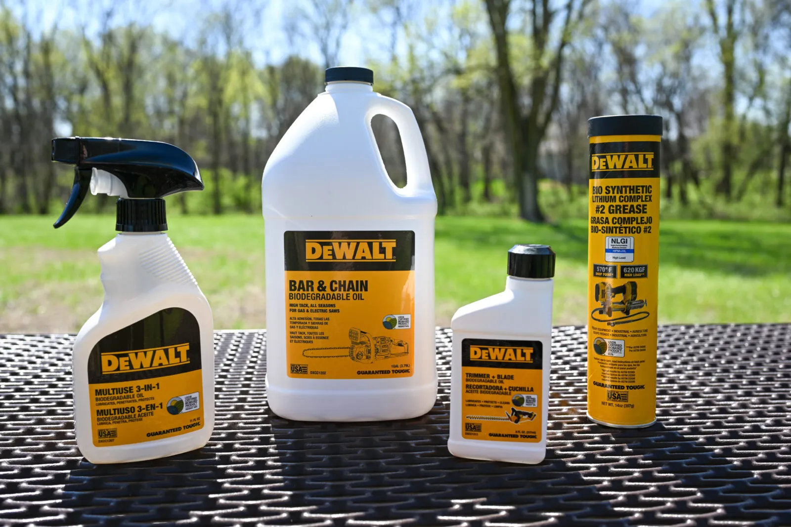 Four DEWALT biodegradable maintenance products displayed outdoors on a metal table: a multiuse 3-in-1 oil spray bottle, a large jug of biodegradable bar and chain oil, a small bottle of trimmer and blade oil, and a can of bio-synthetic lithium grease, with green trees and grass in the background.
