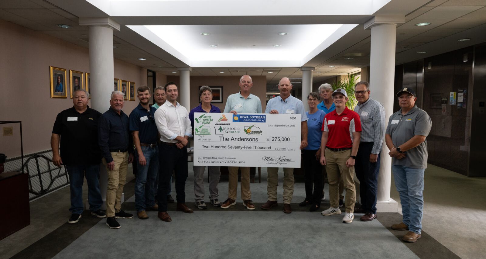 A group of men and women standing indoors in a lobby-like space, posing together while holding an oversized ceremonial check, with framed photos on the walls behind them.