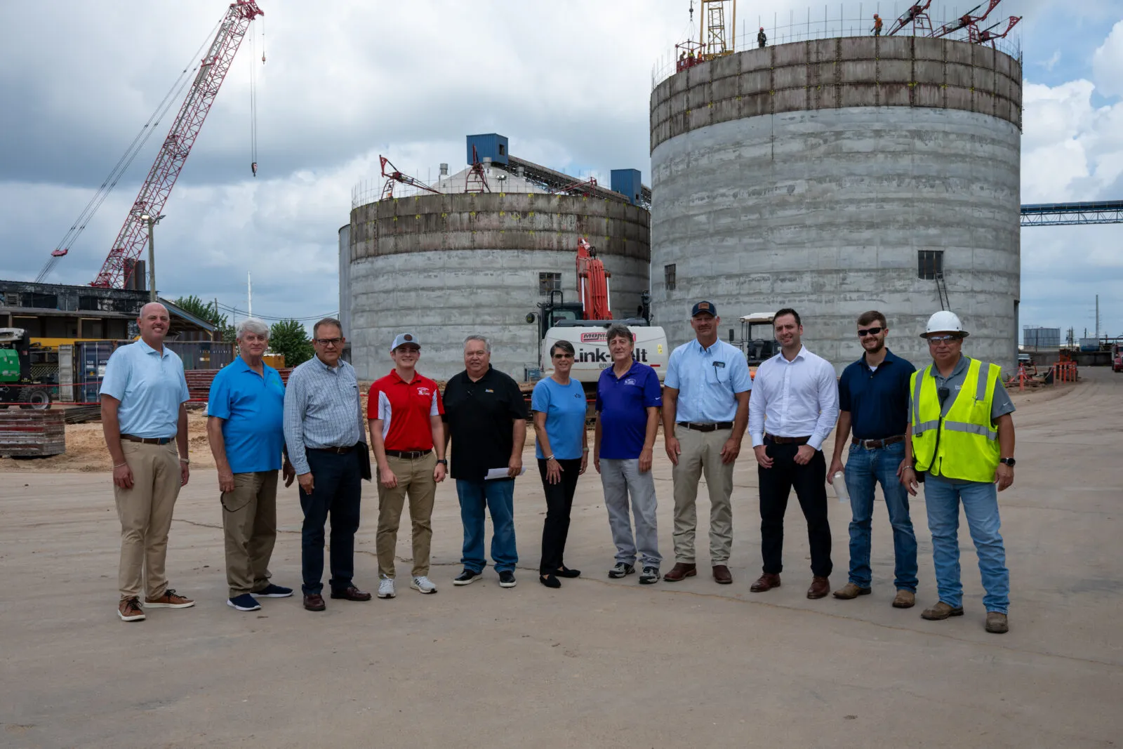 A group of eleven people standing on a construction site in front of two large cylindrical concrete storage tanks under construction, with cranes, heavy equipment, and scaffolding visible in the background beneath a cloudy sky.