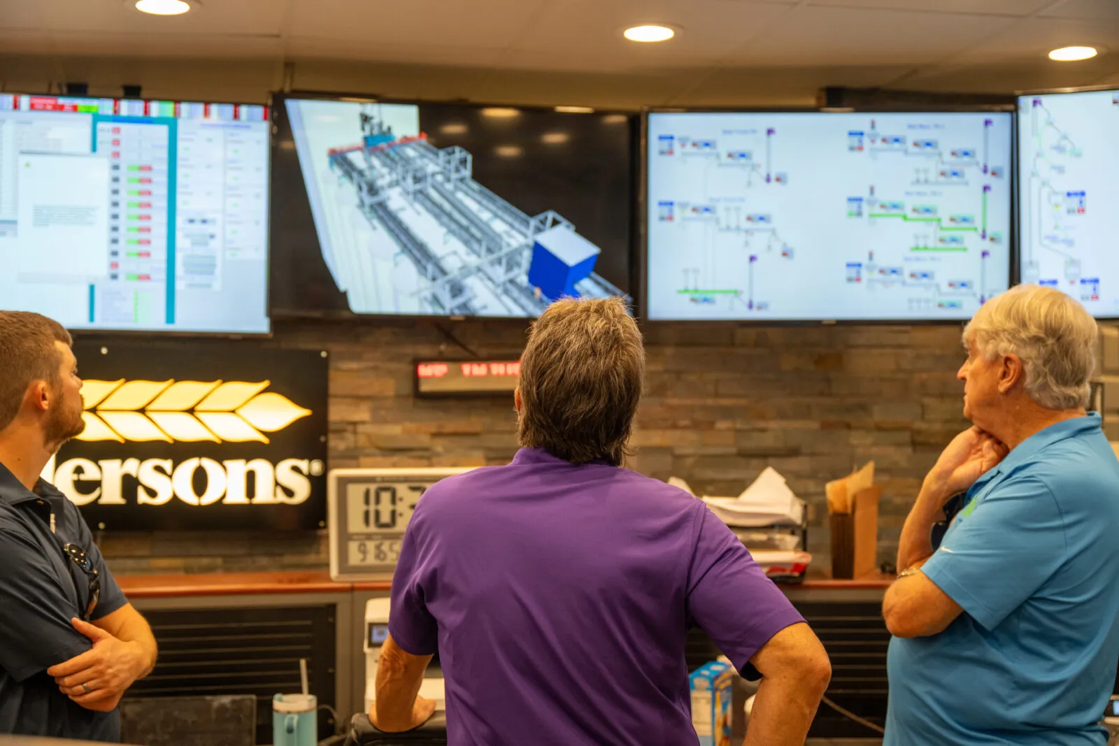 Three people standing in a control room, viewed from behind, watching multiple large monitors that display industrial process diagrams and a 3D facility model above a workstation with a digital clock and equipment.