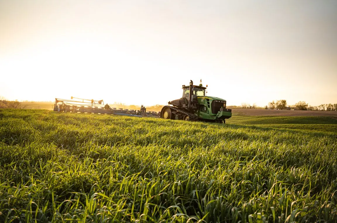 A tractor pulling farm equipment across a lush green field at sunset, with rolling farmland in the background.