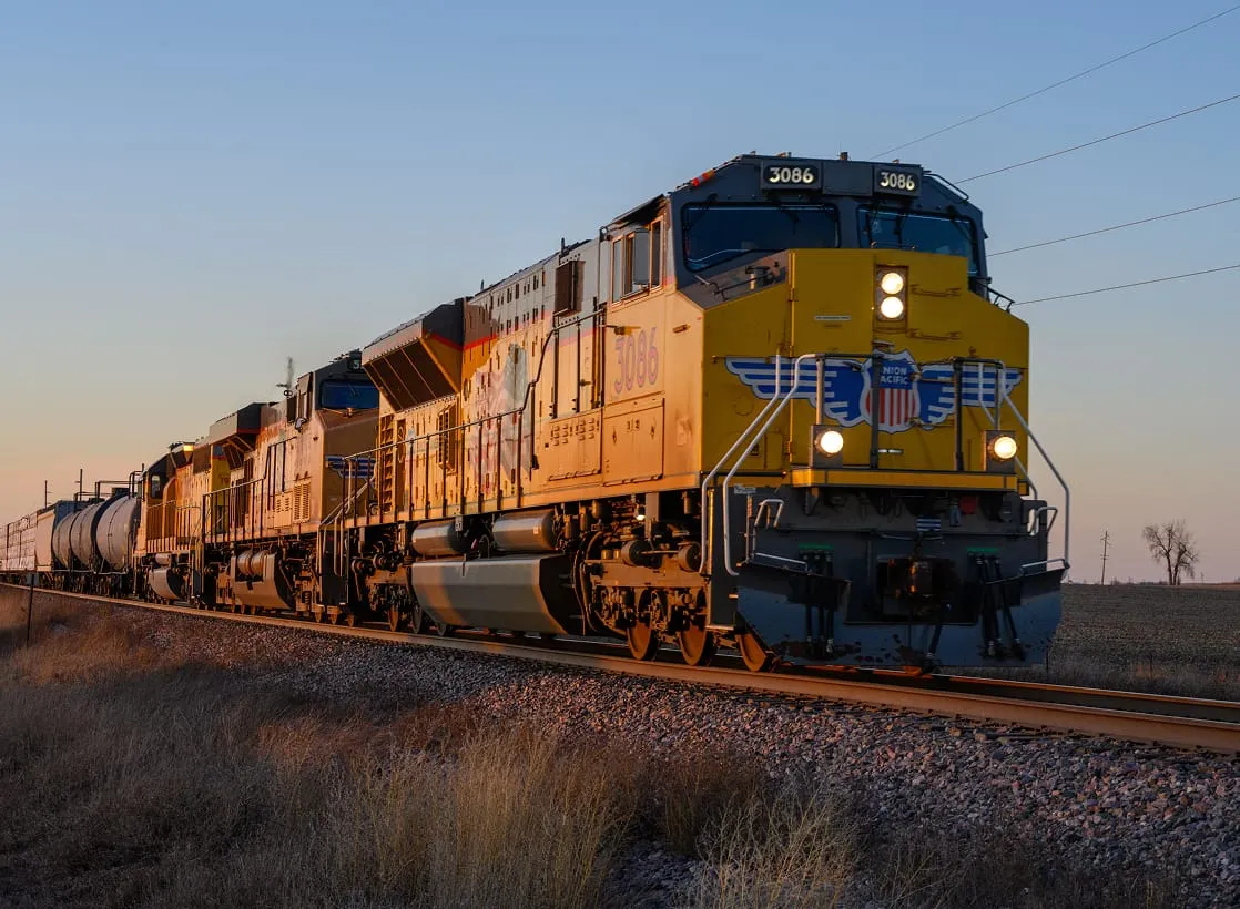 A powerful yellow freight train pulls tank cars along a rural track at sunset.