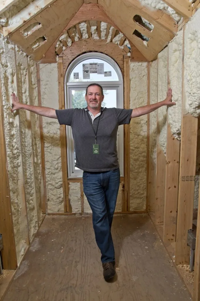A man standing inside a house under renovation, surrounded by newly installed spray foam insulation on the walls.