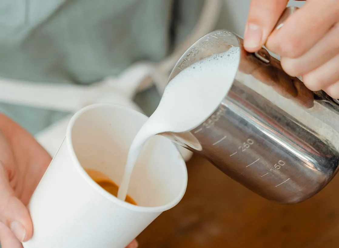 Steamed milk is being poured from a metal pitcher into a cup of espresso.