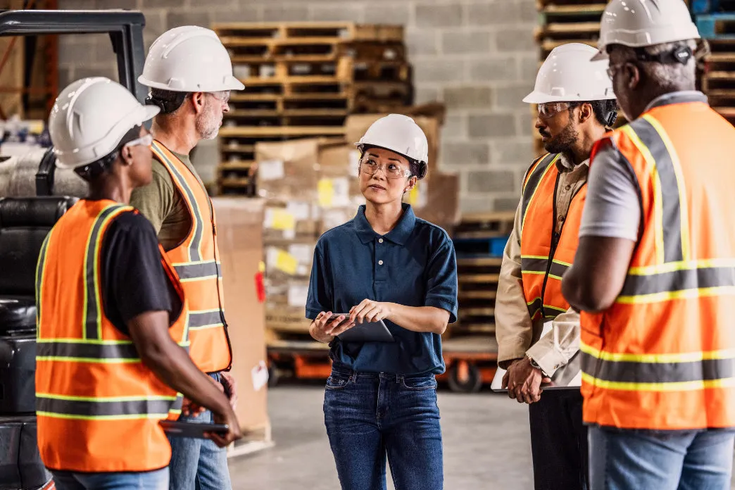 A group of warehouse workers wearing safety helmets and reflective vests listen attentively to a consultant holding a tablet, who appears to be providing guidance in an industrial setting.