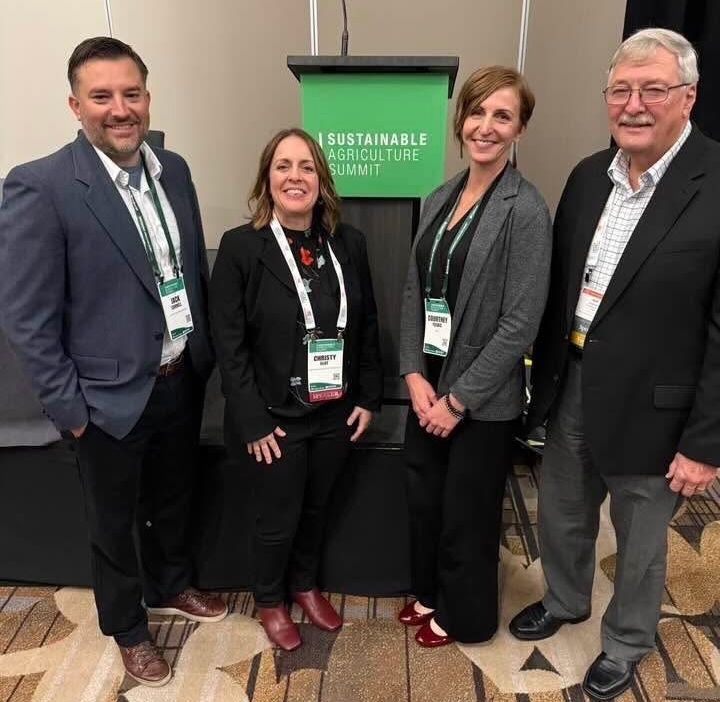 Four people standing together in front of a podium with a green ‘Sustainable Agriculture Summit’ sign, posing for a group photo at a conference.