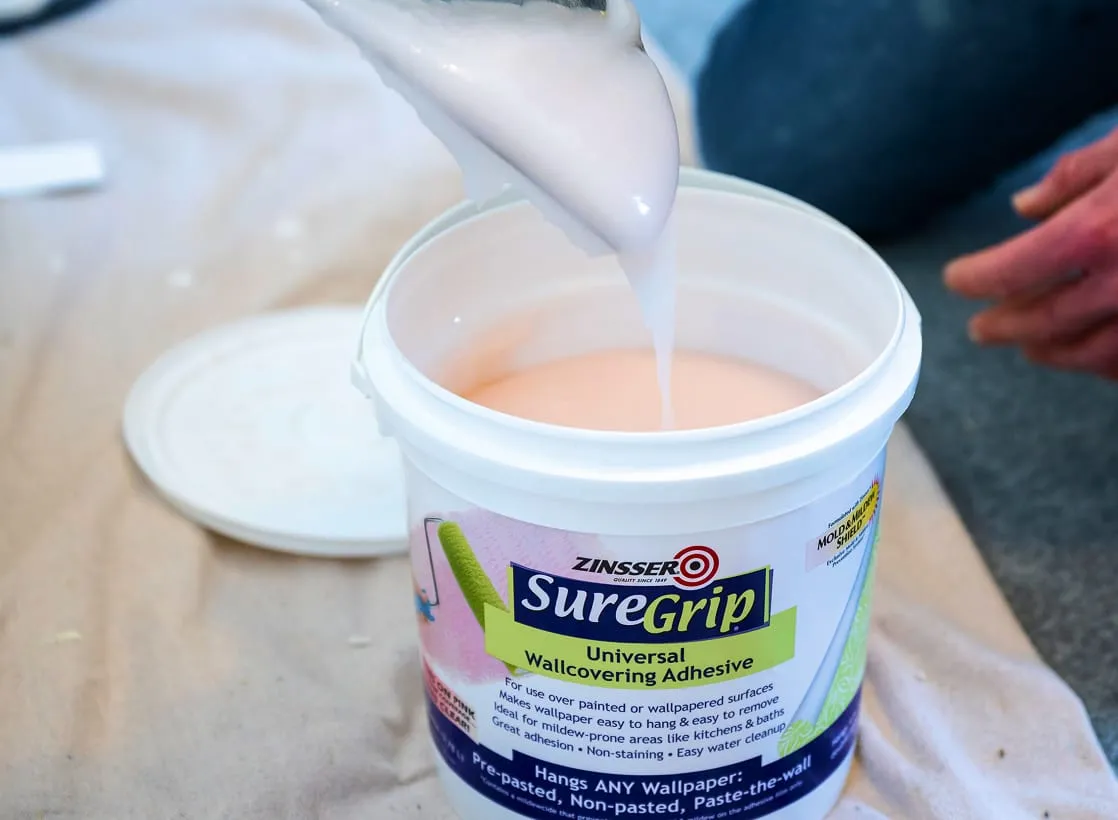 A container of wallpaper adhesive sits open as pale paste drips from a tool above it.