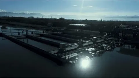 Aerial view of a fish or aquaculture facility with rectangular tanks and walkways, sunlight reflecting off the water, and mountains in the background.