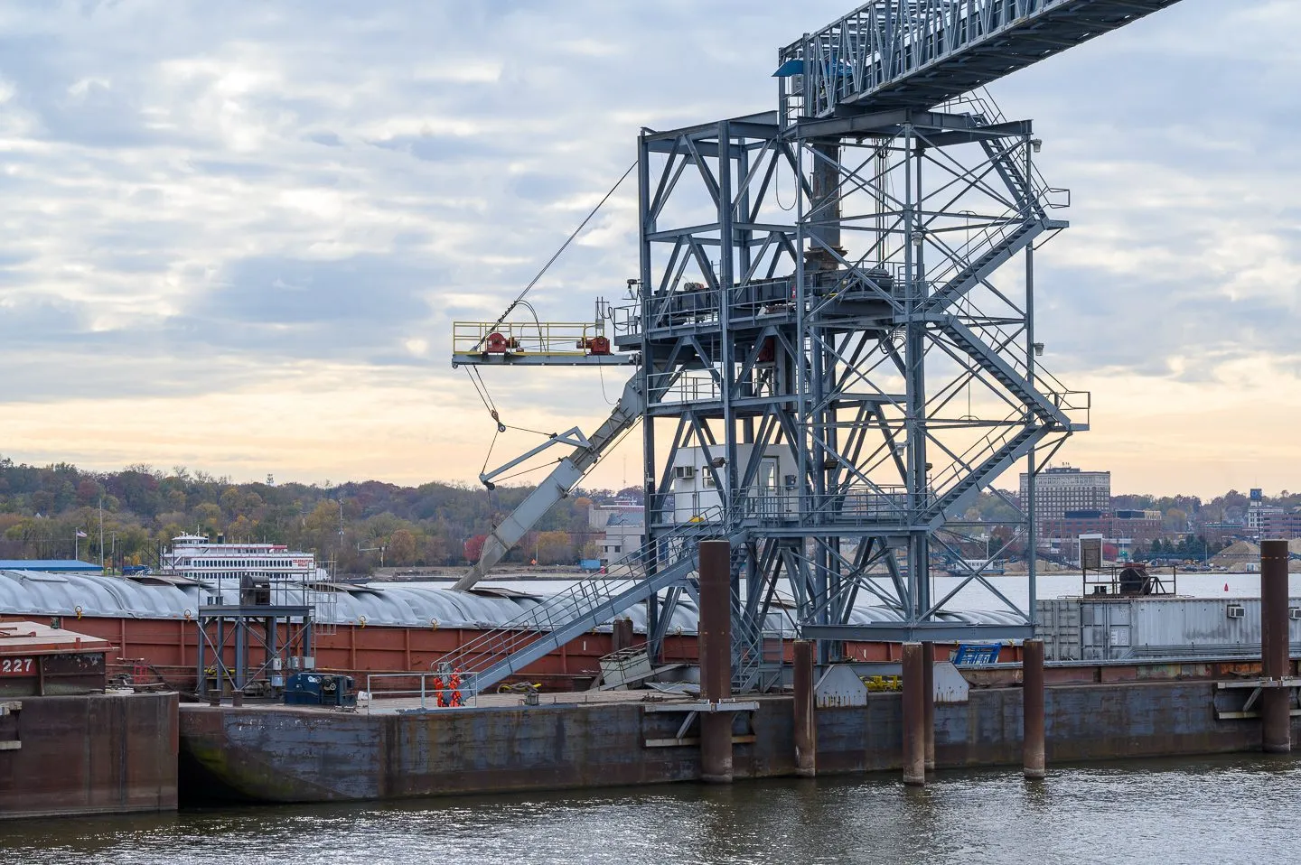 Soybeans being loaded onto a barge beneath a large industrial conveyor structure on a river.