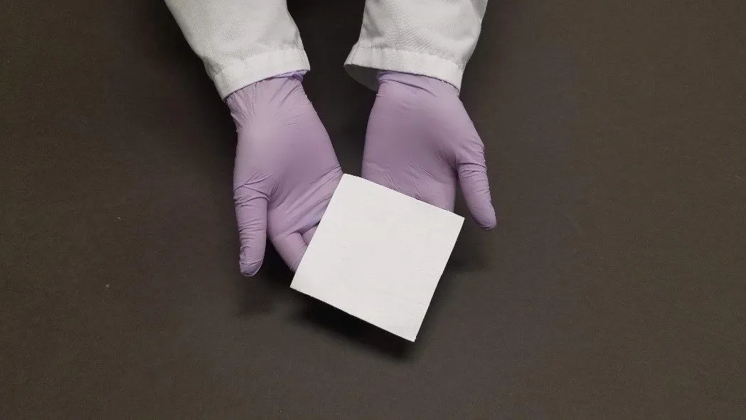 Gloved hands holding a small square white material sample against a dark background.