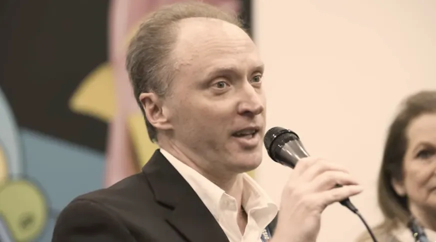 A man speaking into a microphone during an event, with colorful artwork blurred in the background.”