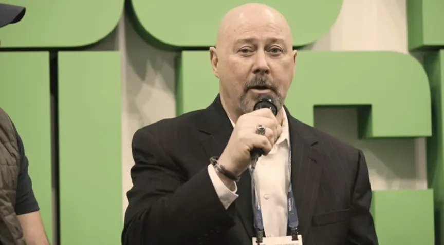 A man in a dark suit speaking into a microphone on a stage with a green backdrop.